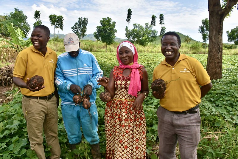 two Tanzanian sweet potato farmers stand in a field smiling and holding up sweet potatoes with two Feed the Future staff