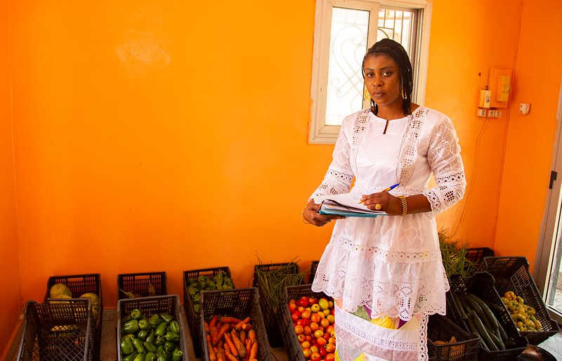 food aid worker holding paperwork while standing in front of baskets of fruits and vegetables