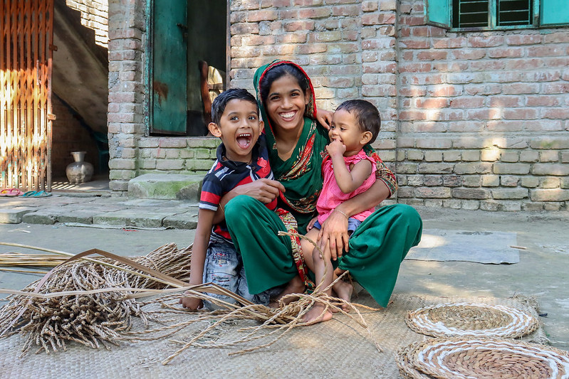 Young mother, boy, and girl sit on the ground smiling next to a weaving project